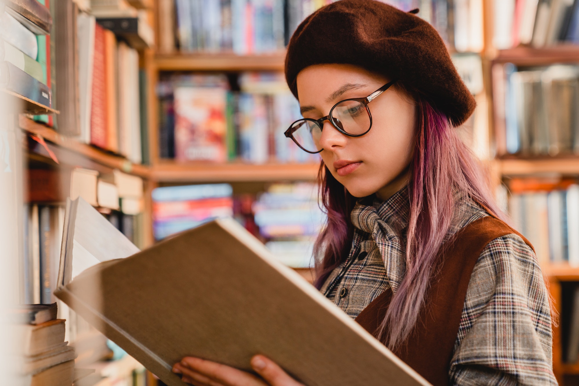 Intelligent young caucasian girl in glasses reading a book at the library